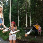 Eine Frau hilft einem Kind beim Überqueren einer Baumstammschaukel in einem Wald, während ein anderer Erwachsener und ein Kind von einer Bank im Hintergrund zusehen.