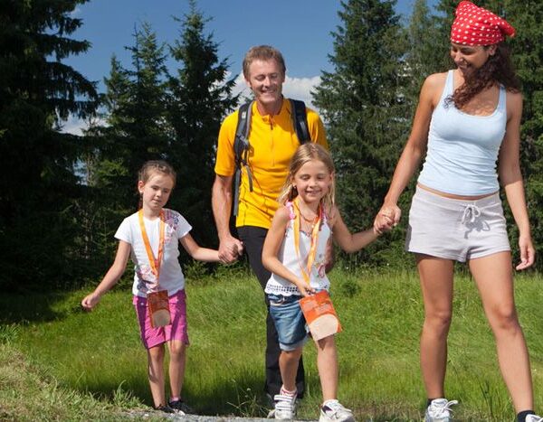 Eine vierköpfige Familie, zwei Erwachsene und zwei Kinder, geht an einem sonnigen Tag gemeinsam auf einem Graspfad in einem Waldgebiet spazieren.
