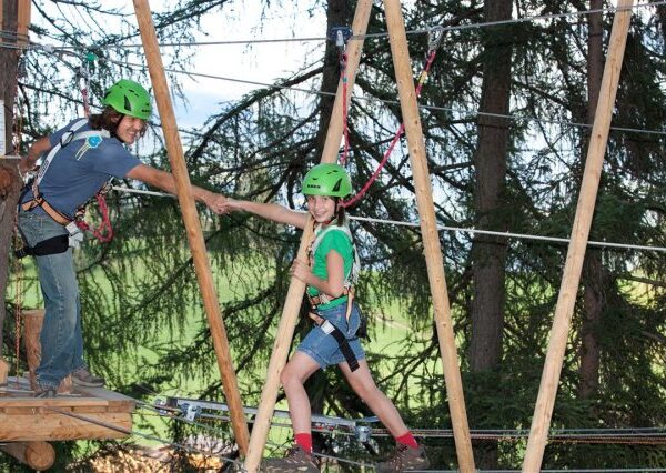 Zwei Personen mit Helmen und Gurten geben sich auf einem Hochseilgarten zwischen Bäumen die Hand, an dessen Struktur Sicherheitsleinen befestigt sind.