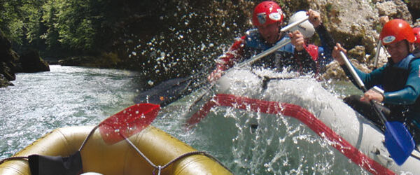Eine Gruppe von Menschen mit Helmen und Schwimmwesten paddelt auf einem Wildwasserfloß durch einen Fluss und spritzt dabei Wasser auf, während sie durch die Stromschnellen navigieren.