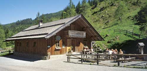 Eine Holzhütte mit Balkon steht in einer grasbewachsenen Hügellandschaft; draußen sitzen an einem sonnigen Tag mehrere Personen an Picknicktischen.