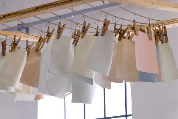 Papierbögen in verschiedenen Pastellfarben hängen an einem hölzernen Gestell mit Wäscheklammern, das in einem Raum in der Nähe großer Fenster angebracht ist.