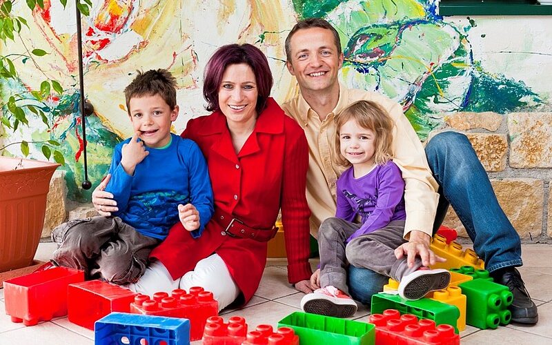 Eine vierköpfige Familie sitzt auf dem Boden, umgeben von großen bunten Spielzeugklötzen, und lächelt in einem hell dekorierten Innenraum in die Kamera.