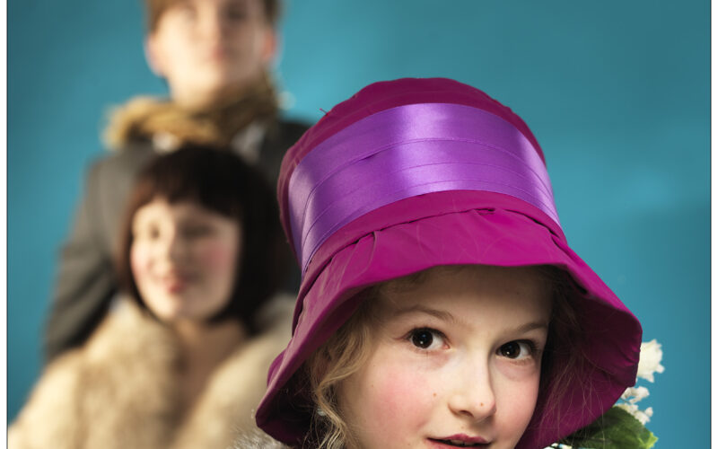 Im Fokus steht ein junges Mädchen mit einem leuchtend lila Hut und einer Pelzstola, im Hintergrund sind vor einem blauen Hintergrund zwei Personen unscharf zu sehen.