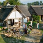 Menschen in historischen Kostümen versammeln sich um strohgedeckte Gebäude und widmen sich in einer rekonstruierten Dorfkulisse Outdoor-Aktivitäten und Kunsthandwerk.