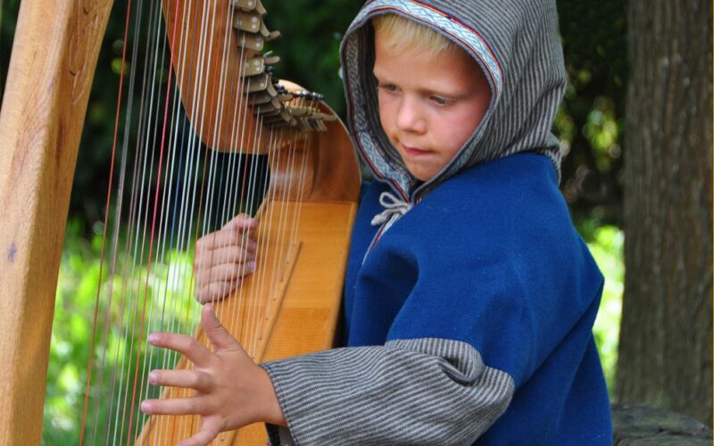 Ein kleines Kind in einem blauen Kapuzenmantel spielt im Freien eine Holzharfe. Dabei konzentriert es sich mit einer Hand auf die Saiten und hält mit der anderen das Instrument.