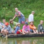 Eine Gruppe Kinder sitzt und steht auf einem Holzfloß, das auf einem Teich schwimmt, umgeben von grüner Vegetation.