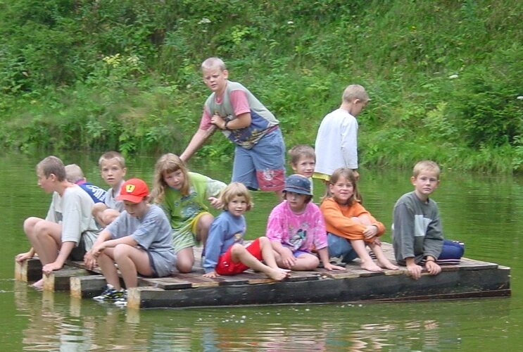 Eine Gruppe Kinder sitzt und steht auf einem Holzfloß, das auf einem Teich schwimmt, umgeben von grüner Vegetation.