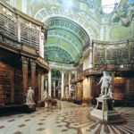 Große, verzierte Bibliothek mit hohen, gewölbten Decken, detaillierten Fresken, hohen Bücherregalen, Marmorstatuen und einem gemusterten Fußboden.