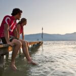 Zwei Menschen sitzen mit den Füßen im Wasser auf einem Holzsteg und beobachten den Sonnenuntergang über einem See mit Bergen im Hintergrund.