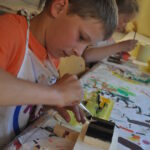 Ein Junge in einer Schürze bemalt mit einem Pinsel eine Holzarbeit an einem Tisch, der mit bunten Farbspritzern bedeckt ist. Ein anderes Kind arbeitet im Hintergrund.