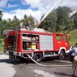 Ein rotes Feuerwehrauto steht mit ausgefahrenem Schlauch auf einer Straße; zwei Personen stehen darauf und eine weitere Person fotografiert die Szene.