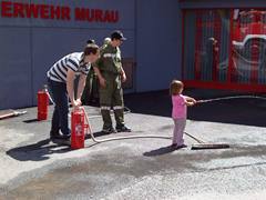 Ein Kind sprüht Wasser aus einem Schlauch, unterstützt von zwei Erwachsenen, in der Nähe von Feuerlöschern vor einem Gebäude mit der Aufschrift „FEUERWEHR MURAU“.