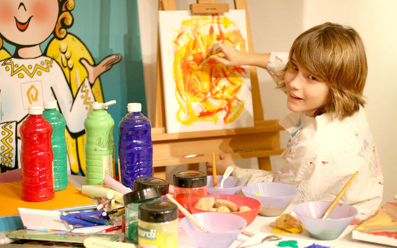 Ein Kind malt mit gelben und roten Farben auf einer Leinwand an einem Tisch voller Künstlerbedarf, darunter Farbflaschen, Pinsel und Schüsseln.