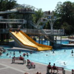Menschen schwimmen in einem öffentlichen Schwimmbad und nutzen eine große gelbe Wasserrutsche, während andere von Bänken und schattigen Bereichen in der Nähe aus zuschauen.