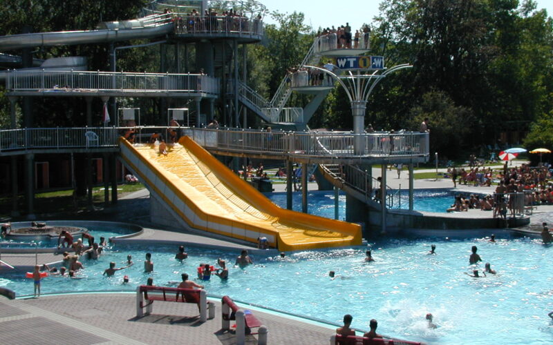 Menschen schwimmen in einem öffentlichen Schwimmbad und nutzen eine große gelbe Wasserrutsche, während andere von Bänken und schattigen Bereichen in der Nähe aus zuschauen.
