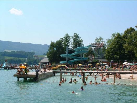 Überfüllter Badebereich am Seeufer mit Menschen an einem Sandstrand, im Wasser und auf einem Pier; im Hintergrund sind eine große blaue Wasserrutsche und Bäume zu sehen.