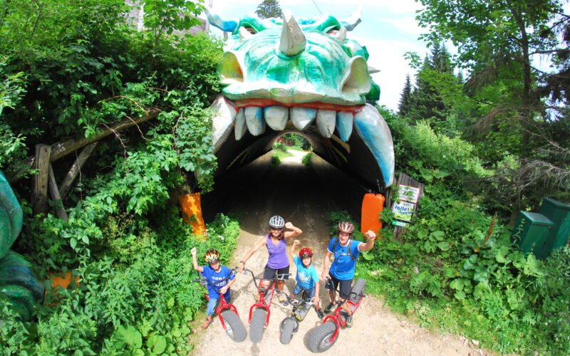 Vier Kinder mit Fahrrädern und Helmen posieren vor einem Tunneleingang, der wie ein Drachenmaul aussieht, umgeben von Grünpflanzen.