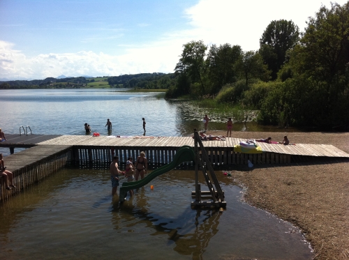 Kinder spielen auf einer Rutsche im flachen Wasser in der Nähe eines hölzernen Stegs an einem Strand am See, während sich im Hintergrund Menschen entspannen und schwimmen.