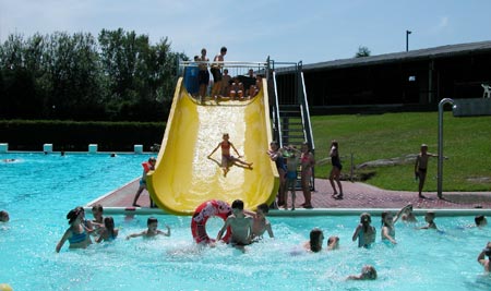 Kinder spielen in einem Freibad, einige schwimmen, während andere eine gelbe Wasserrutsche benutzen; die Szene ist lebhaft und spielt sich an einem sonnigen Tag ab.