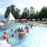 Menschen verschiedenen Alters schwimmen und entspannen sich in einem Außenpool mit Sonnenschirmen, Bäumen und Bergen im Hintergrund.