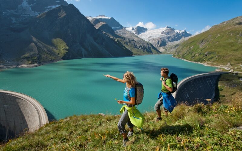 Zwei Wanderer mit Rucksäcken stehen auf einem grasbewachsenen Hang mit Blick auf einen türkisfarbenen See, der von Bergen und einem großen Staudamm begrenzt wird, unter einem klaren blauen Himmel.