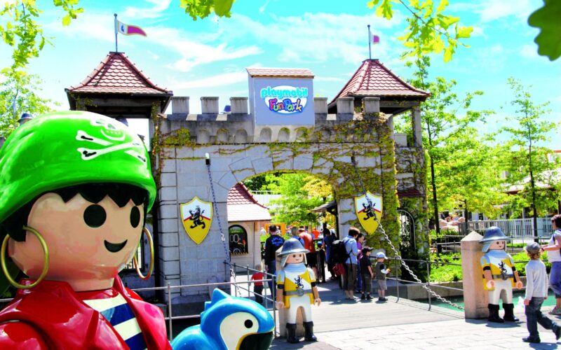 Eingang zum Playmobil FunPark mit großen Playmobil-Figurenstatuen, einem burgähnlichen Tor und Besuchern, die an einem sonnigen Tag eintreten.
