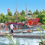 Menschen laufen auf Holzbrücken und Flößen über einen Teich in einem Park im Freien mit einem großen roten Piratenschiff und grünen Bäumen im Hintergrund.