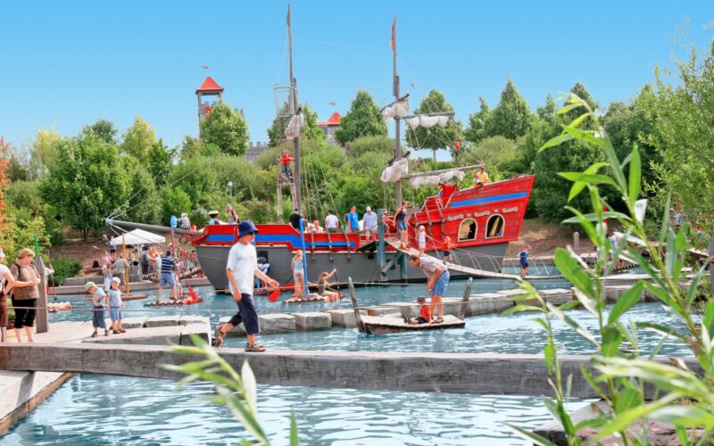 Menschen laufen auf Holzbrücken und Flößen über einen Teich in einem Park im Freien mit einem großen roten Piratenschiff und grünen Bäumen im Hintergrund.