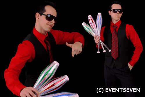 Ein Mann in rotem Hemd, schwarzer Weste, gestreifter Krawatte und Sonnenbrille hält vier Jonglierkeulen vor einem schwarzen Hintergrund.