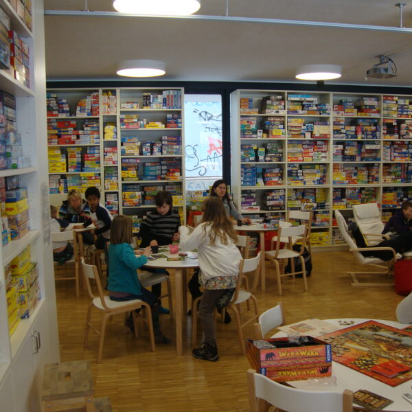 Menschen sitzen an Tischen und spielen Brettspiele in einem hell erleuchteten Raum, der mit Regalen voller verschiedener Brettspiele gefüllt ist.