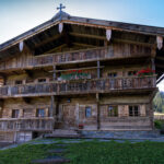 Ein rustikales Alpenchalet aus Holz mit Balkonen, Blumenkästen und einem Kreuz auf dem Dach, eingebettet in eine grasbewachsene Berglandschaft unter einem klaren blauen Himmel.