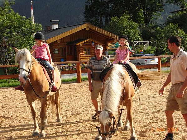 Zwei kleine Kinder mit Helmen sitzen auf Pferden, jeweils in Begleitung eines Erwachsenen, auf einem Reitplatz in der Nähe einer Blockhütte.