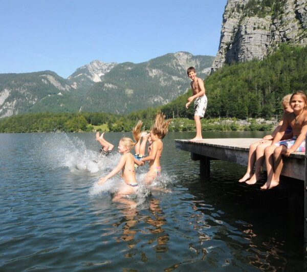 Mehrere Kinder springen von einem hölzernen Steg in einen See, während ein Junge steht und ein Mädchen auf dem Steg sitzt; im Hintergrund sind Berge und Bäume zu sehen.