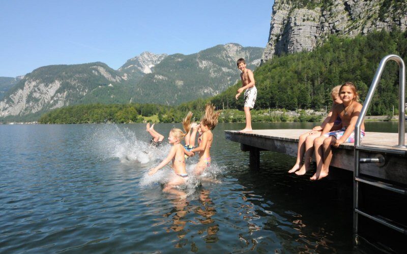 Mehrere Kinder springen von einem hölzernen Steg in einen See, während ein Junge steht und ein Mädchen auf dem Steg sitzt; im Hintergrund sind Berge und Bäume zu sehen.