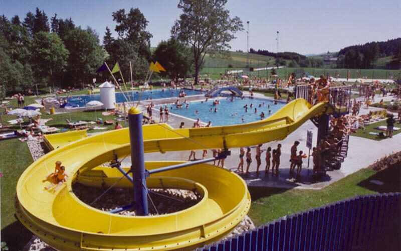 Öffentliches Freibad mit einer großen gelben Wasserrutsche, umgeben von Menschen, die sich auf einer Wiese sonnen, schwimmen und entspannen.