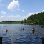 Menschen, die in einem von Bäumen umgebenen See schwimmen und sich entspannen, mit einem Steg und bewölktem Himmel im Hintergrund.