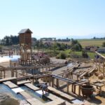 Spielplatz im Freien mit Holzkonstruktionen, Wasserspielbereichen, einem Turm und spielenden Kindern, mit Häusern und Feldern im Hintergrund unter einem klaren Himmel.
