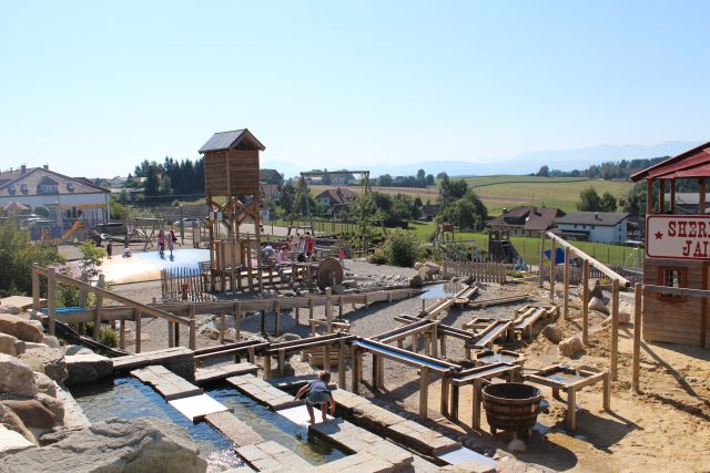 Spielplatz im Freien mit Holzkonstruktionen, Wasserspielbereichen, einem Turm und spielenden Kindern, mit Häusern und Feldern im Hintergrund unter einem klaren Himmel.