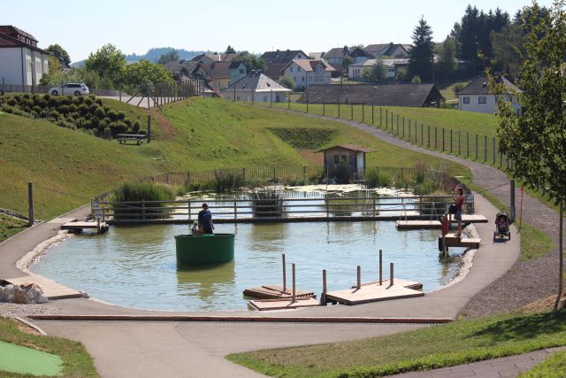 Ein kleiner Teich mit hölzernen Stegen und einer Person in einem runden Boot; ein Kind und ein Kinderwagen sind auf einem Weg zu sehen, mit Häusern und grasbewachsenen Hügeln im Hintergrund.