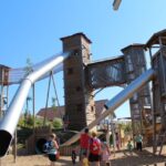 An einem sonnigen Tag erkunden Kinder und Erwachsene eine große hölzerne Spielplatzstruktur mit hohen Türmen, langen Metallrutschen und Seilbrücken.