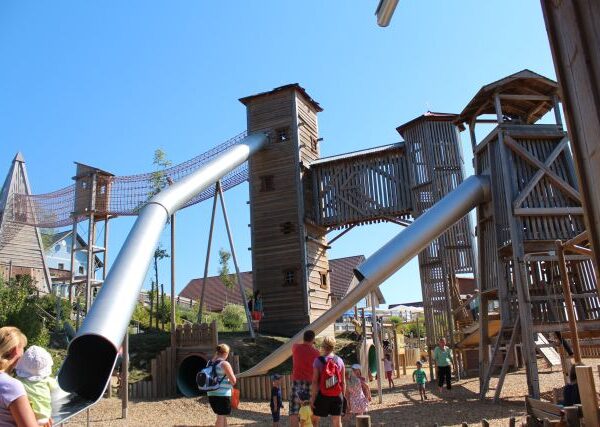 An einem sonnigen Tag erkunden Kinder und Erwachsene eine große hölzerne Spielplatzstruktur mit hohen Türmen, langen Metallrutschen und Seilbrücken.