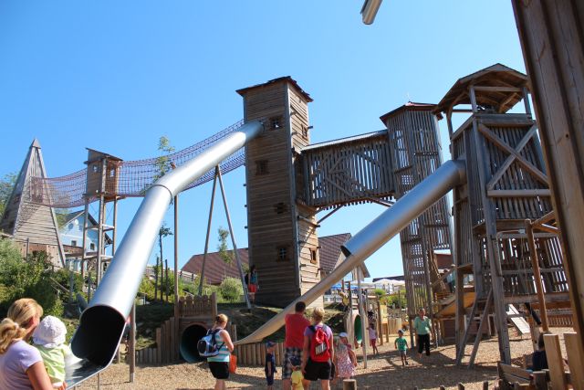 An einem sonnigen Tag erkunden Kinder und Erwachsene eine große hölzerne Spielplatzstruktur mit hohen Türmen, langen Metallrutschen und Seilbrücken.