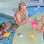 Ein Erwachsener und zwei kleine Kinder spielen mit Plastikspielzeug am Rande eines Schwimmbeckens. Der Erwachsene sitzt im Wasser, während die Kinder einen Eimer und Becher benutzen.