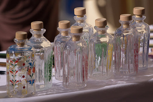 Eine Reihe von Klarglasflaschen mit Holzkorken, einige mit bunten Blumenmustern verziert, auf einer weißen Fläche ausgestellt.