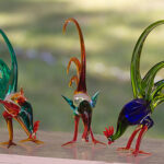 Drei bunte Glashahnfiguren mit langen Schwänzen werden auf einer flachen Oberfläche mit verschwommenem grünem Hintergrund ausgestellt.