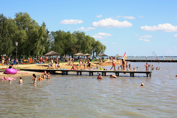 Menschen schwimmen, sonnen sich und entspannen sich an einem Sandstrand am Seeufer mit Bäumen, Sonnenschirmen und einem Holzsteg unter einem teilweise bewölkten Himmel.