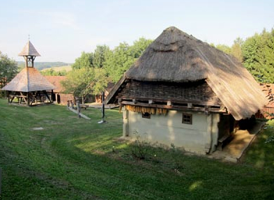 Ein traditionelles Strohdachhaus und eine Holzkonstruktion stehen auf einer Wiese, umgeben von Bäumen und Holzzäunen unter einem klaren Himmel.
