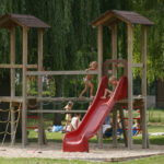 Kinder spielen auf einem hölzernen Spielplatz mit einer roten Rutsche, einer Strickleiter und einer Verbindungsbrücke in einem grasbewachsenen Parkgelände.