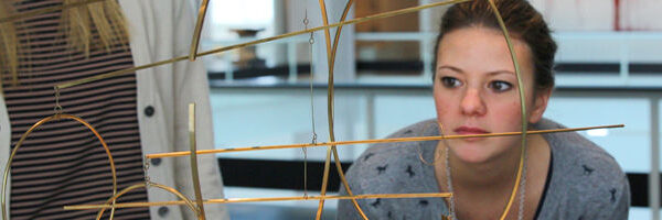 Eine Frau untersucht aufmerksam eine geometrische Metallskulptur, während im Hintergrund eine weitere Person in der Nähe steht.